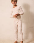 A young boy smiling and wearing a long-sleeve, full-length cream-colored pajama set with a small red star pattern, standing against a warm beige textured wall.