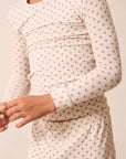 A close-up of a person wearing a long-sleeve cream-colored pajama top with a small repeating red star pattern, paired with matching pajama pants against a plain light beige background.