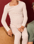A child wearing a long-sleeve crewneck pajama top and matching pants with a small repeating red twinkle star pattern, seated on a tan couch against a dark red wall.