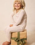A smiling young girl with blonde hair sits barefoot on a brown paper gift box with an olive green ribbon. She is wearing a cream-colored long-sleeve pajama top and matching pants with a small red star pattern.