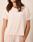 A woman wearing a short-sleeve crewneck pajama top and matching pants in a cream color with a small red star pattern, photographed against a solid beige background.