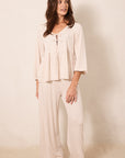 A woman modeling a two-piece pajama set consisting of a long-sleeve peplum top and matching wide-leg pants, both in a cream color with a small red star pattern, against a warm beige studio background.