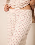 A close-up of a person wearing a matching two-piece sleepwear set with a cream base and small red star pattern. The top is gathered at the waist, and the pants have an elastic waistband.