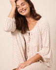 A woman wearing a cream-colored, long-sleeve tie-front top and matching pants with a small red star pattern, posing against a warm beige background.
