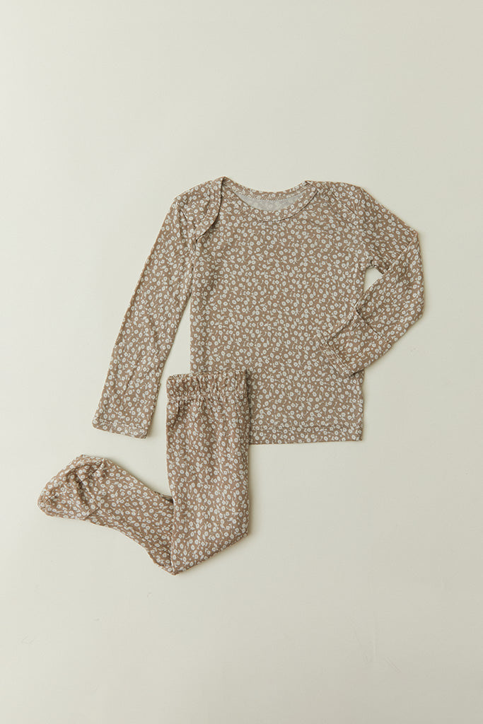 A flat lay of Solly Baby's two-piece baby pajamas in the Cream Floral Print, featuring petite cream buds gently scattered atop a soft taupe brown base. The pajamas feature footed bottoms and an easy wide-neck top and are available in sizes 0m-12m.