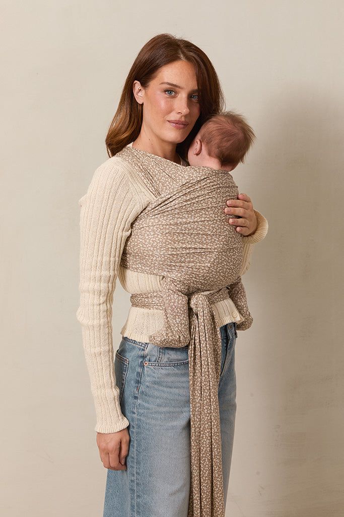 A side view of a woman standing and holding her baby in a Solly Baby Wrap carrier in the Cream Floral print, featuring delicate cream flowers on a taupe background.