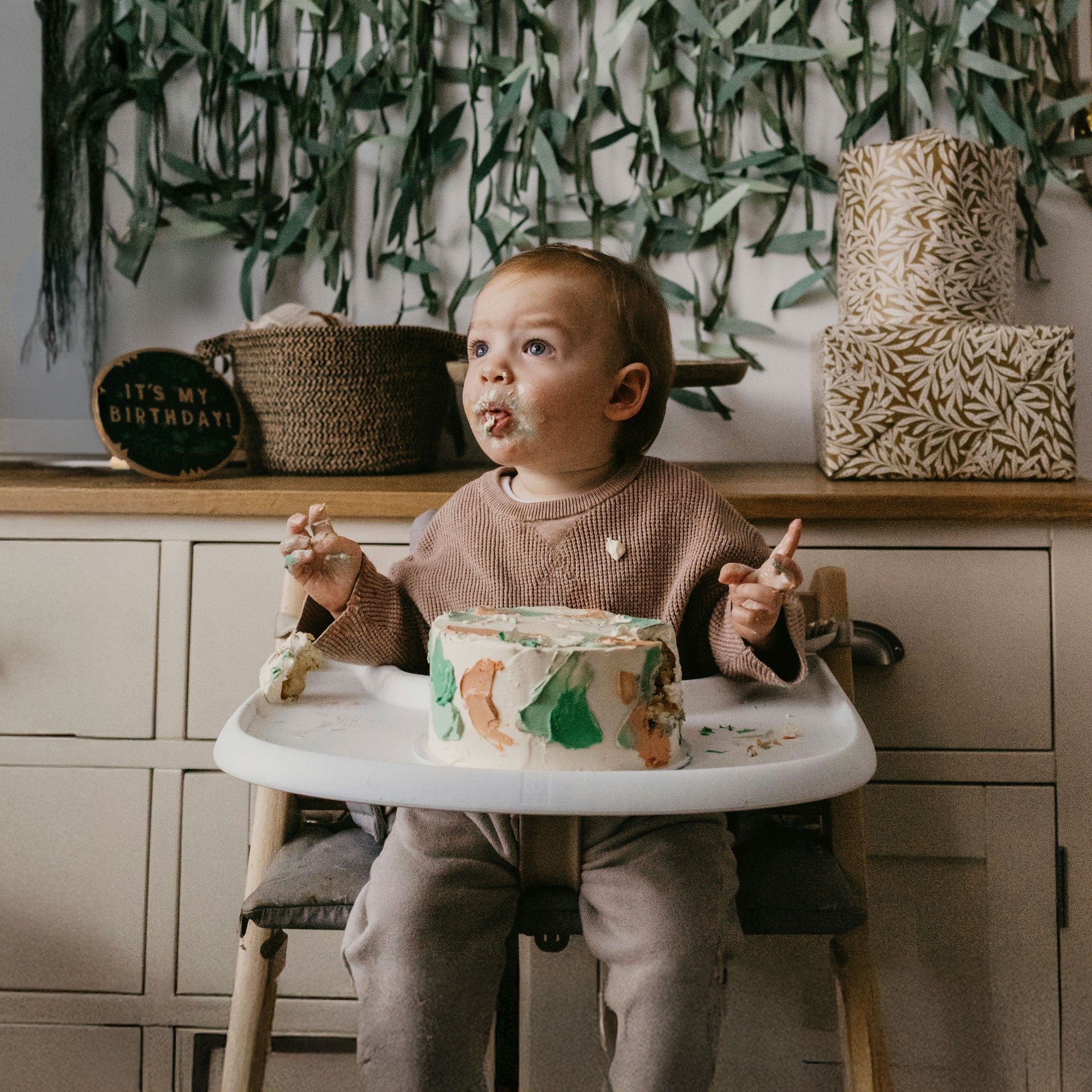 Baby celebrating first birthday with a smash cake in a high chair, surrounded by party decorations and presents.