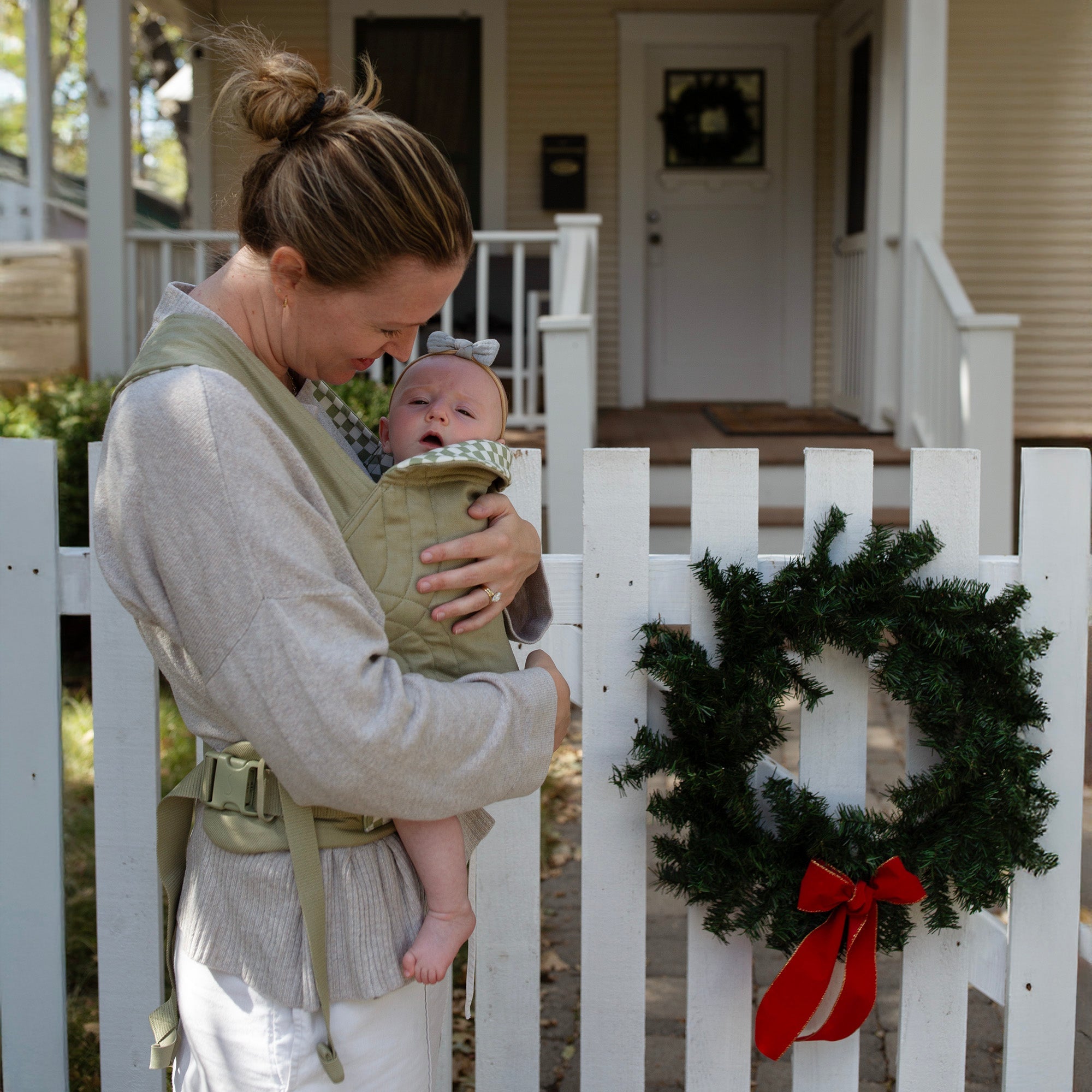 Mother babywears in the Sage Chequer Soft Carrier in front of a white picket fence with a holiday wreath hanging in front of her home