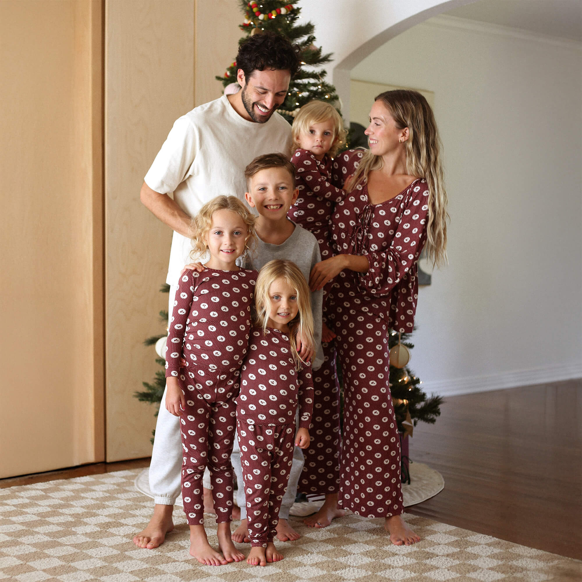 Family of 6 smiles in front of a Christmas tree in a cozy family room wearing matching Solly sleep sets in Pomegranate. 