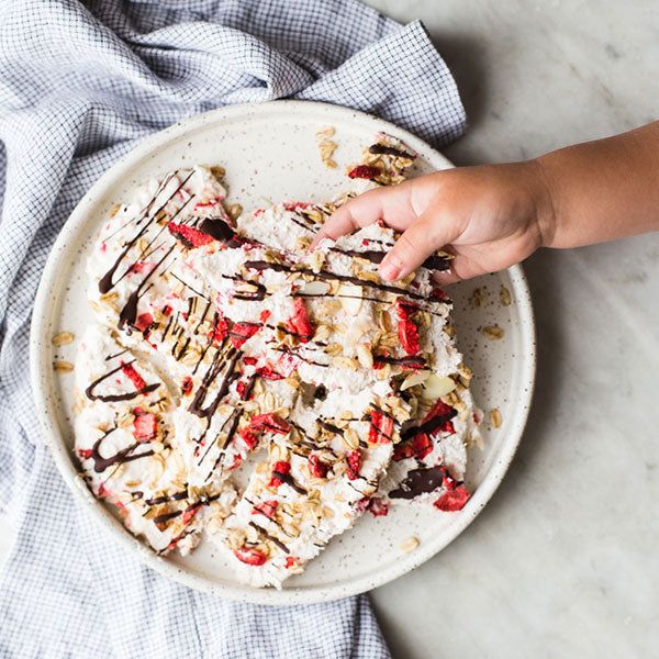 Hand reaching for chocolate drizzled strawberry yogurt bark on a plate on a white marble countertop