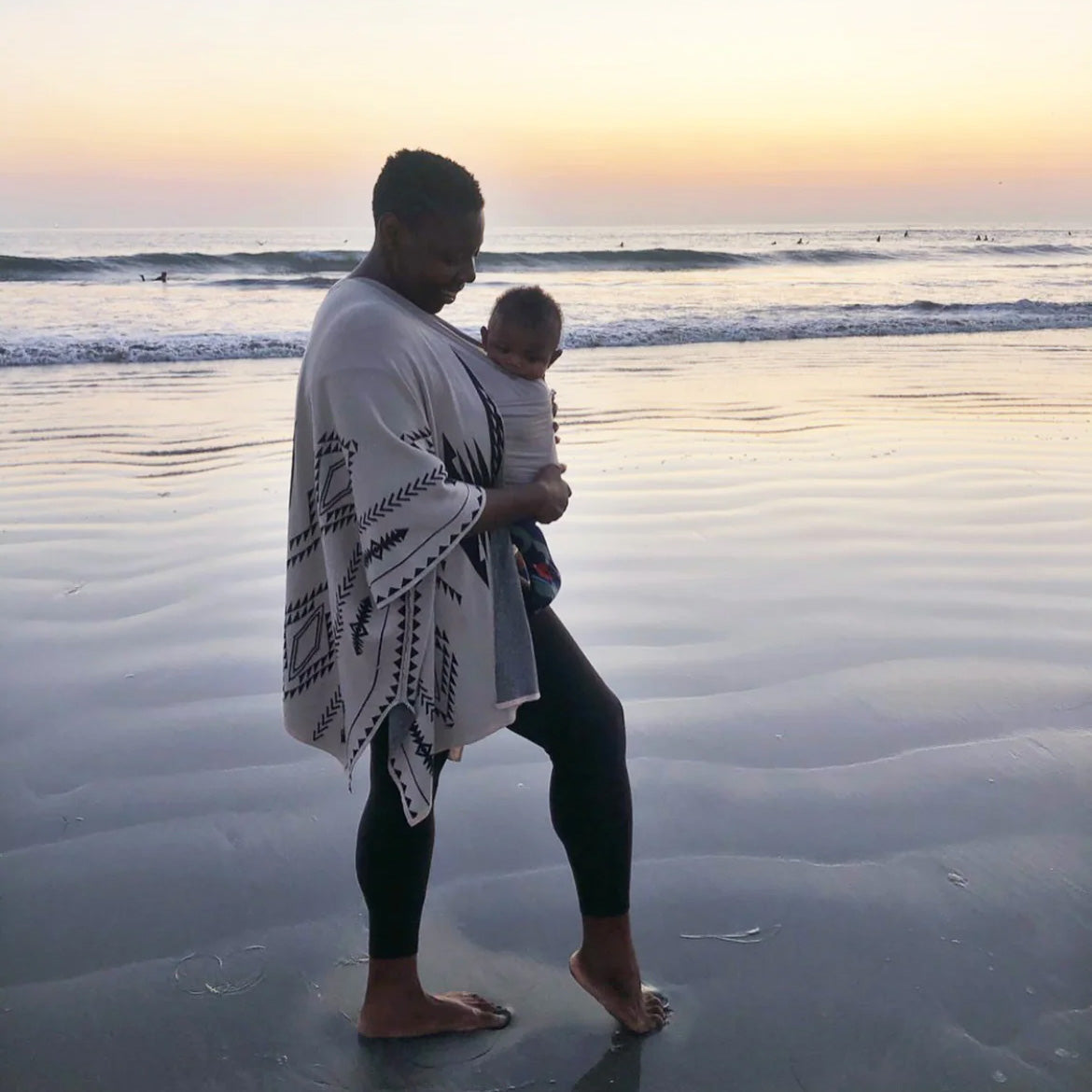 Woman stands on the beach at sunset wearing baby in a Solly Baby wrap