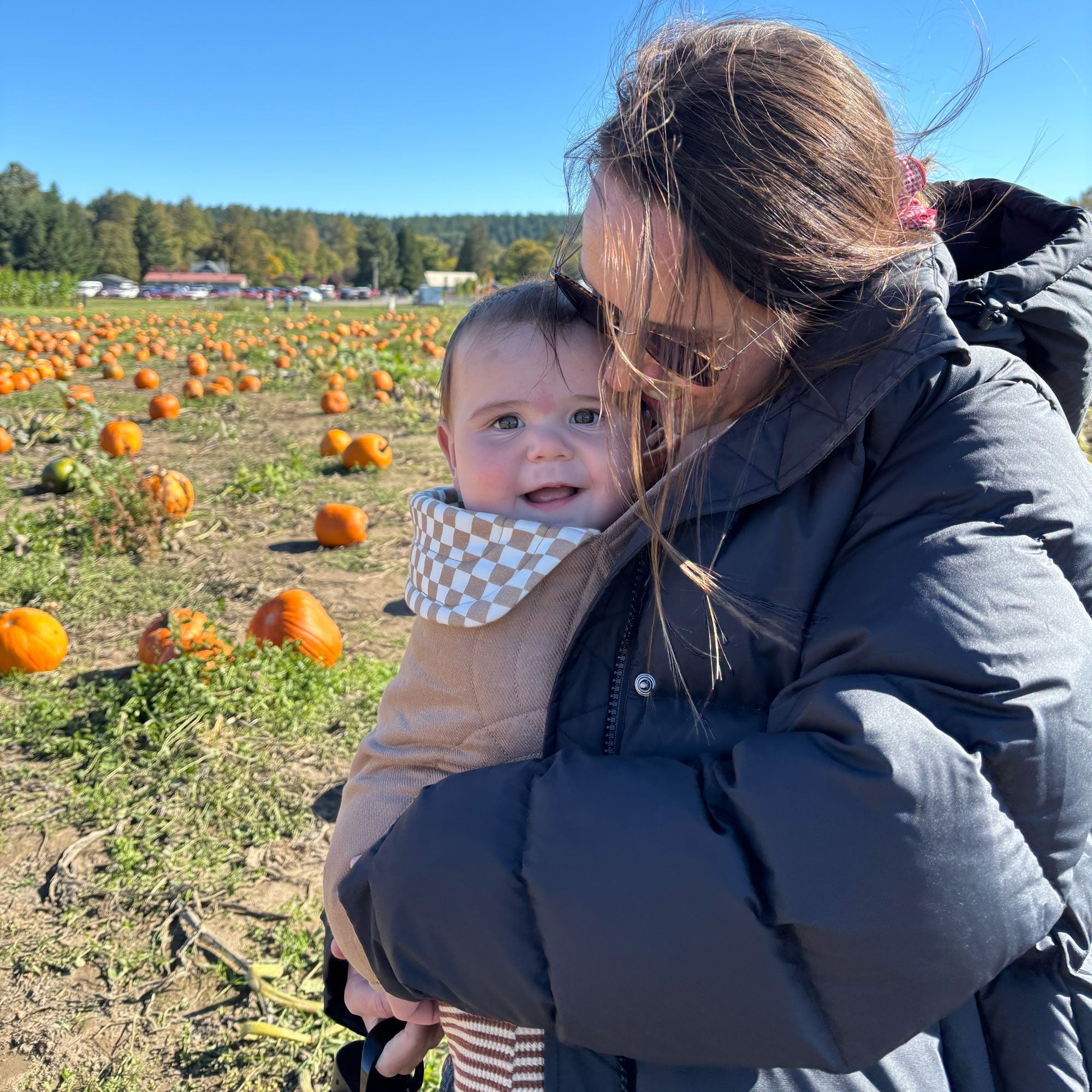 Woman in a black puffer coat stands in a pumpkin patch and looks down at her daughter smiling in the Almondine Chequer Soft Carrier 