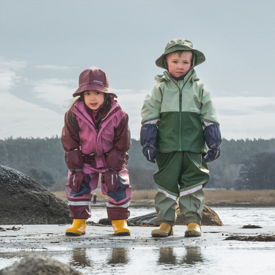 2 children stand on the sandy coast on a cold day, dressed in head to toe rain gear and are ready to play