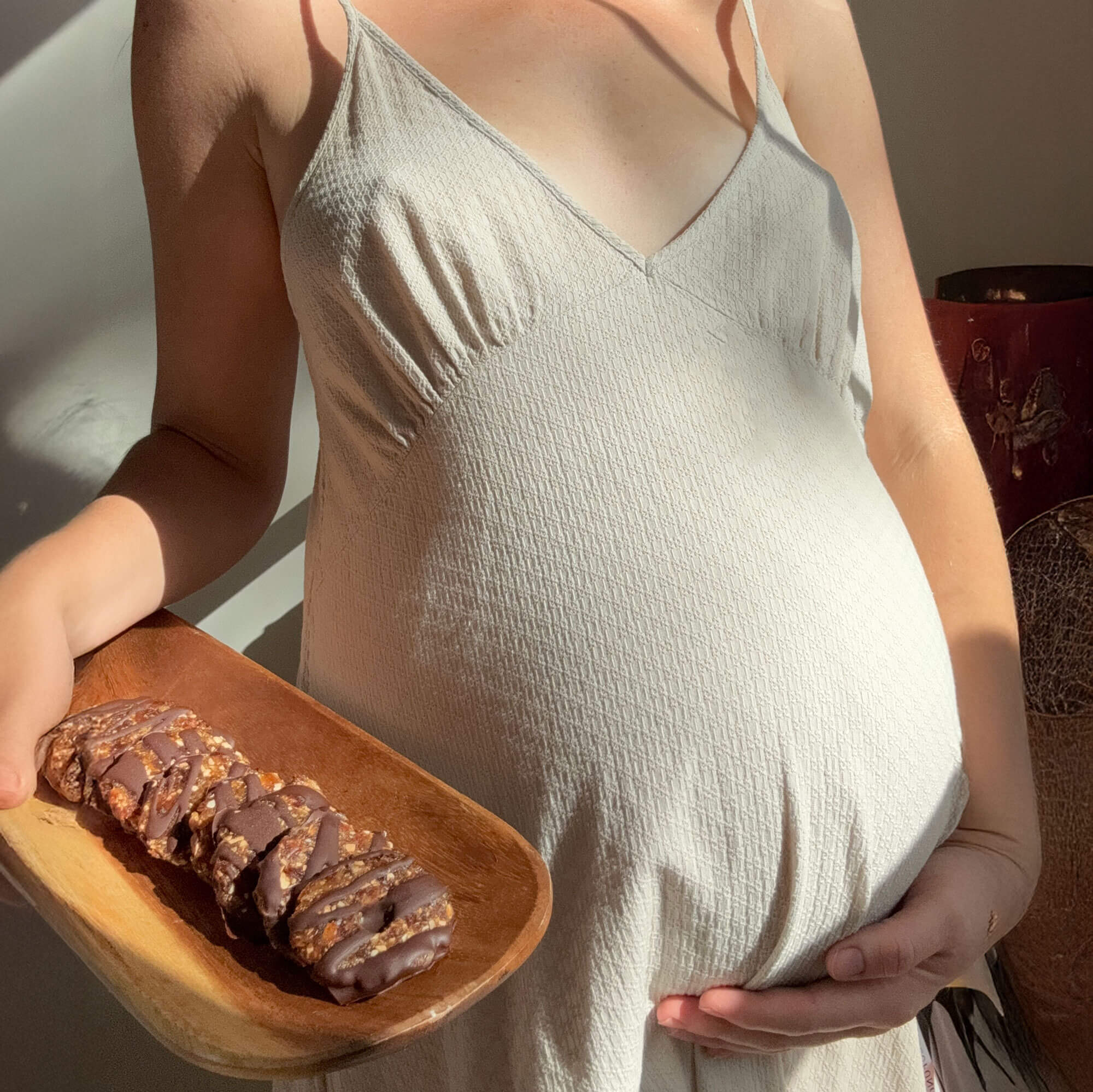 Pregnant woman in neutral dress holding a wooden plate of chocolate-covered date bars to support labor and delivery.