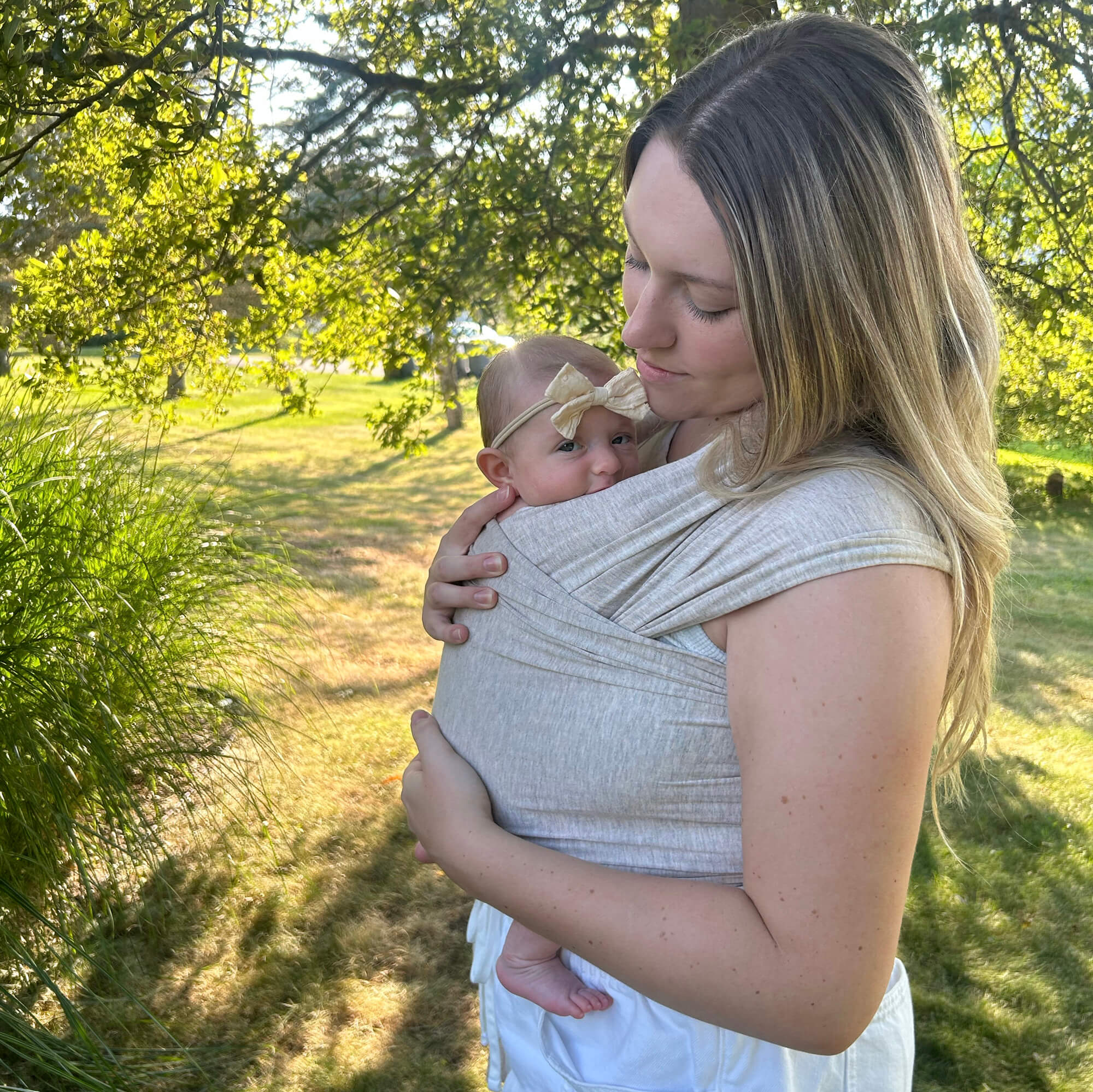 A tender moment of a woman looking lovingly down at her baby in the Flax Solly Wrap. They are outdoors in a sunny grove of trees sharing a moment of connection.