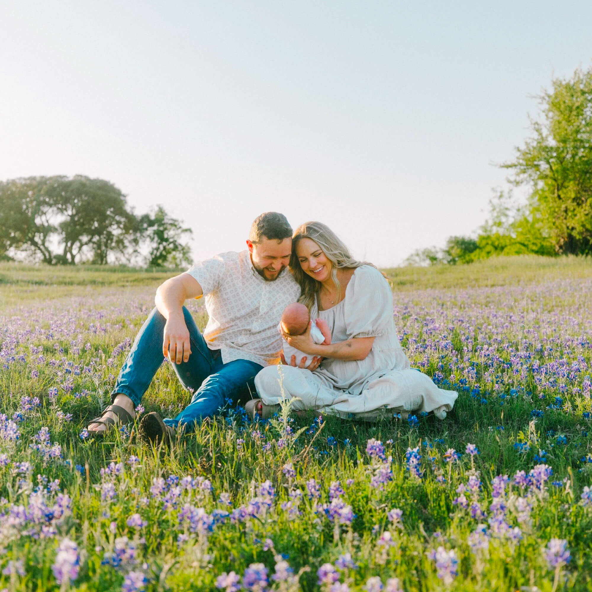 oung family sitting in a blooming wildflower field, smiling and holding their newborn baby.