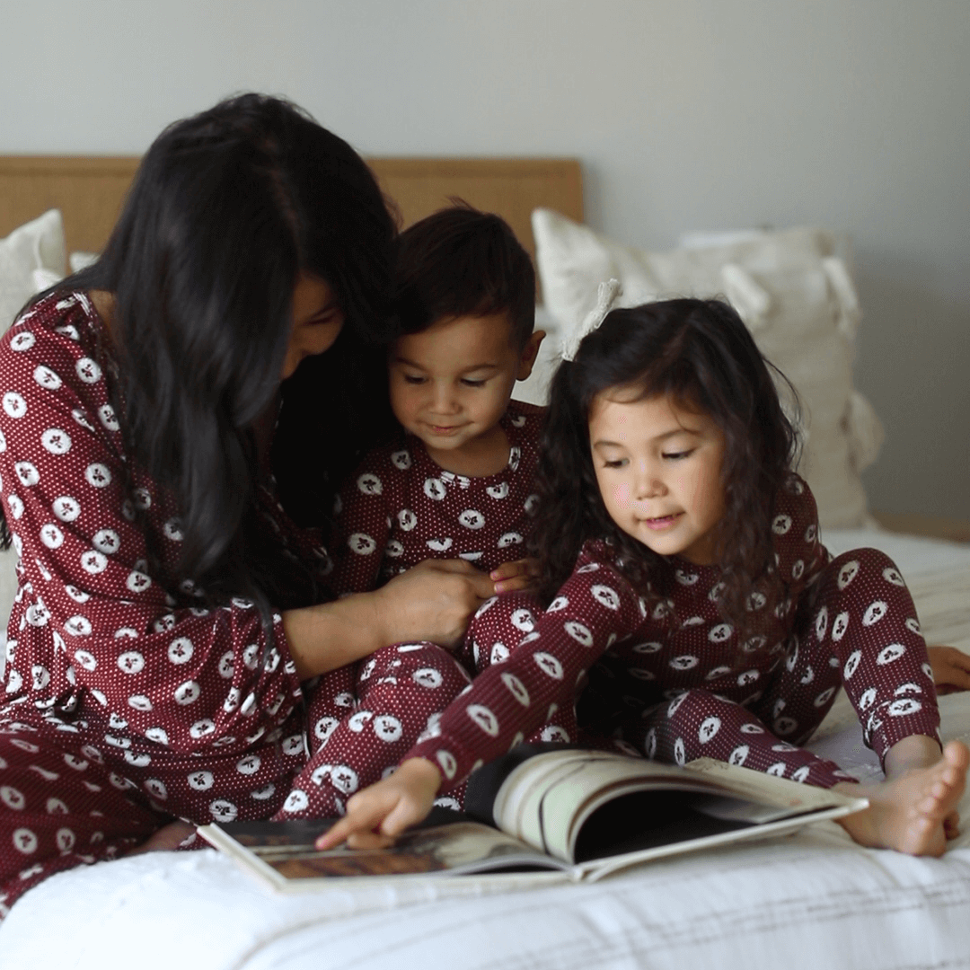Mother and toddler son and daughter sit on a bed in matching Holiday Pajamas from Solly Baby and smile as they read a holiday book together
