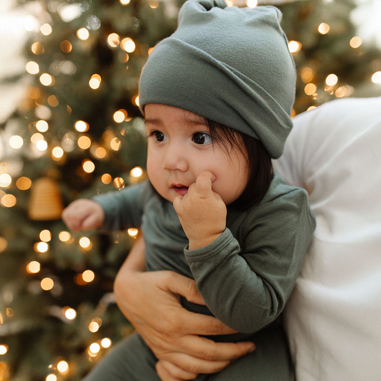 Mother holds baby dressed in the Basil Sleep Set and Knotted Hat in front of the Christmas tree