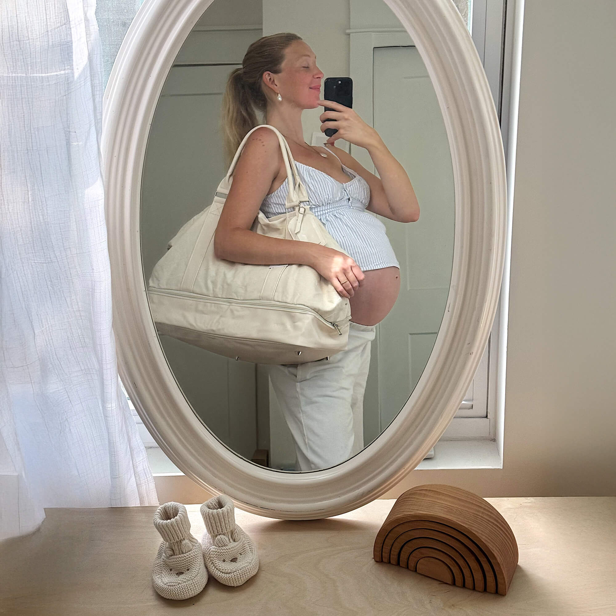 Woman with large, cream hospital bag on shouldertakes a mirror selfie in an oval mirror on a dresser in a baby's nursery
