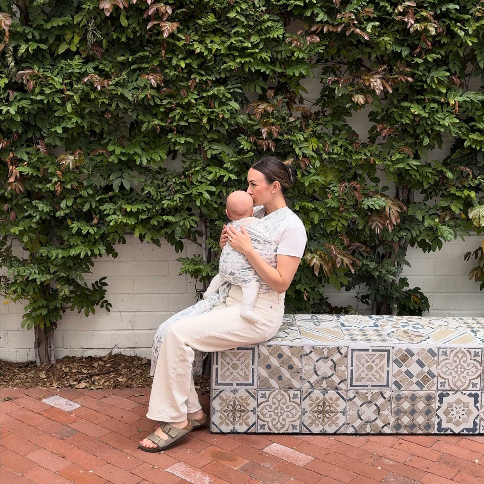 Mom holding baby in Solly Baby Bleu Toile wrap, seated on tiled bench in front of leafy wall.