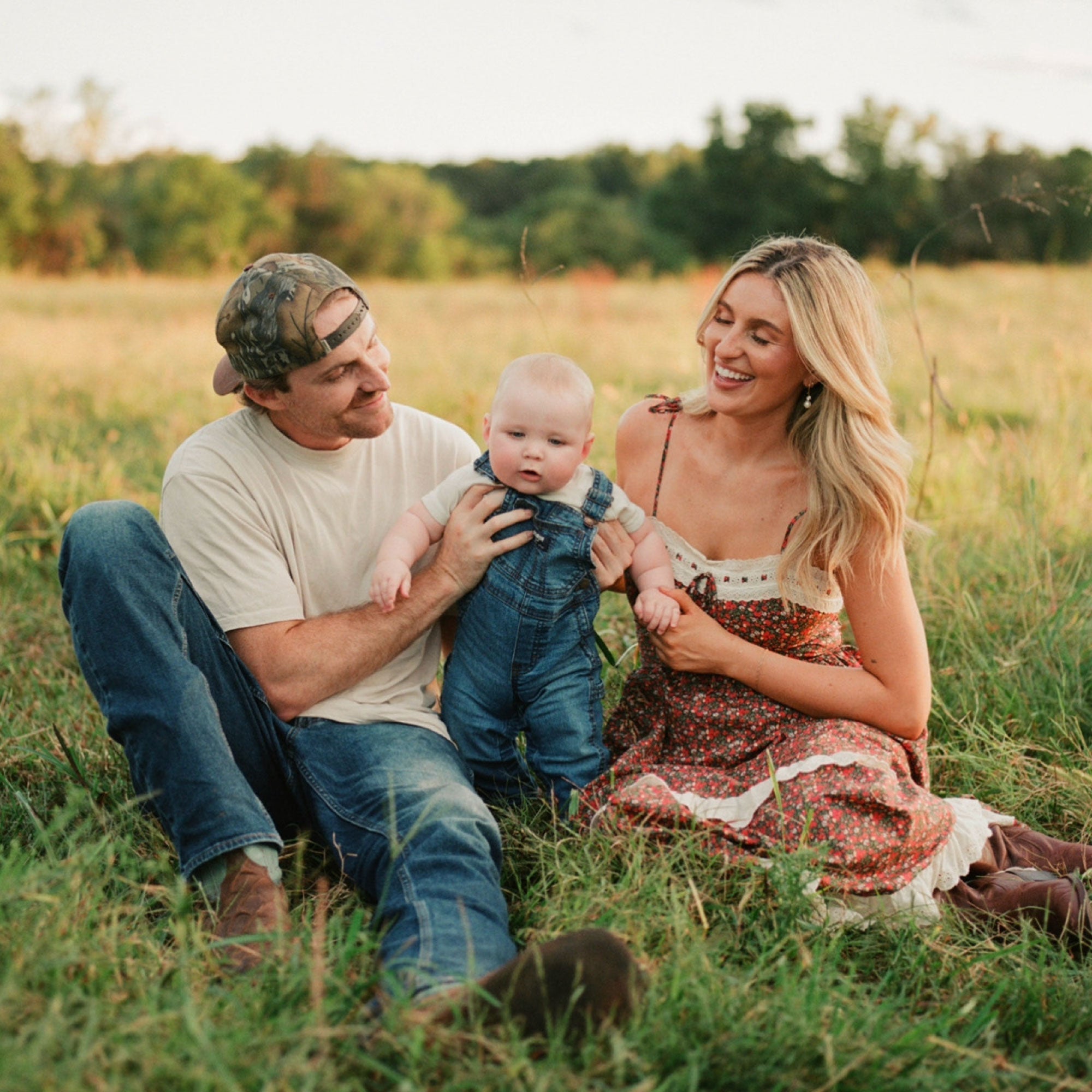Husband and wife sit in a field at golden hour and smile looking down at their baby dressed in denim overalls