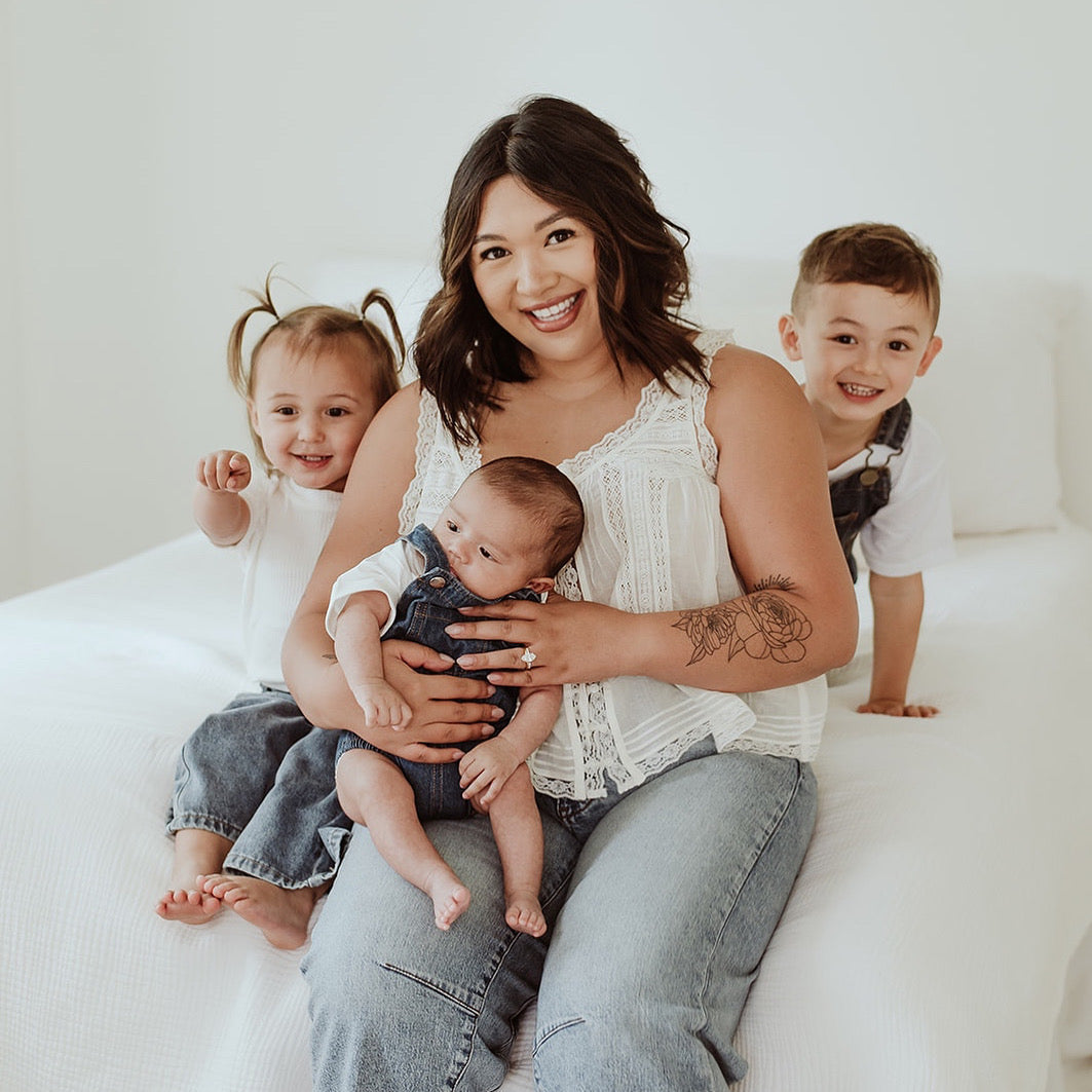 Mom smiling on bed with her three young children, embracing her breastfeeding journey and motherhood.