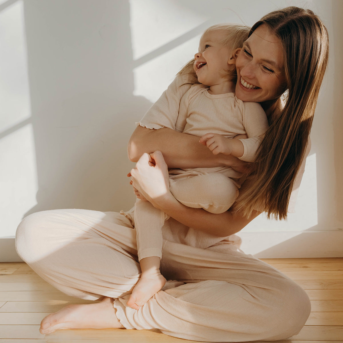 Mom sits cross-legged and holds baby daughter in her arms and both laugh in the light of the window