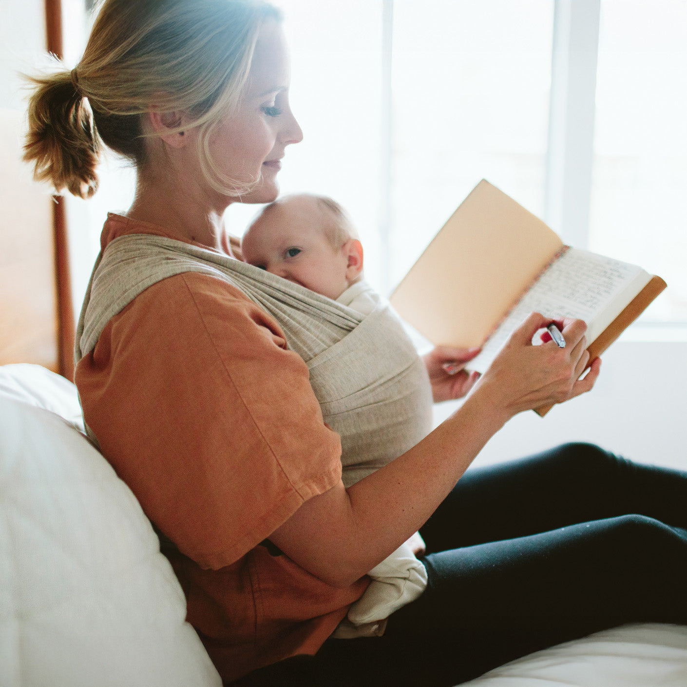 Woman sits comfortably on bed and journals with her baby in the Flax Solly wrap