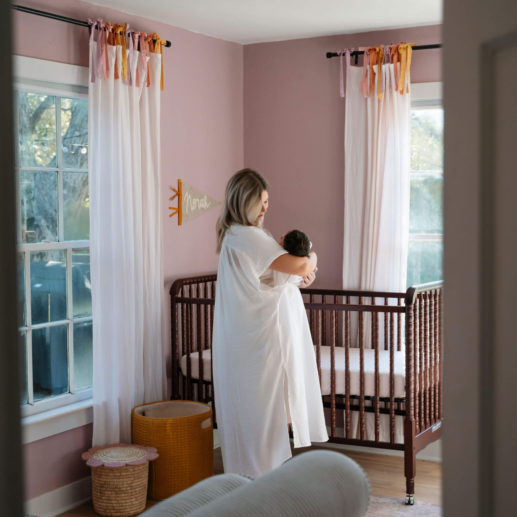 Woman in a white flowy dress holds her baby and looks down lovingly in a pink-walled nursery room with a vintage crib