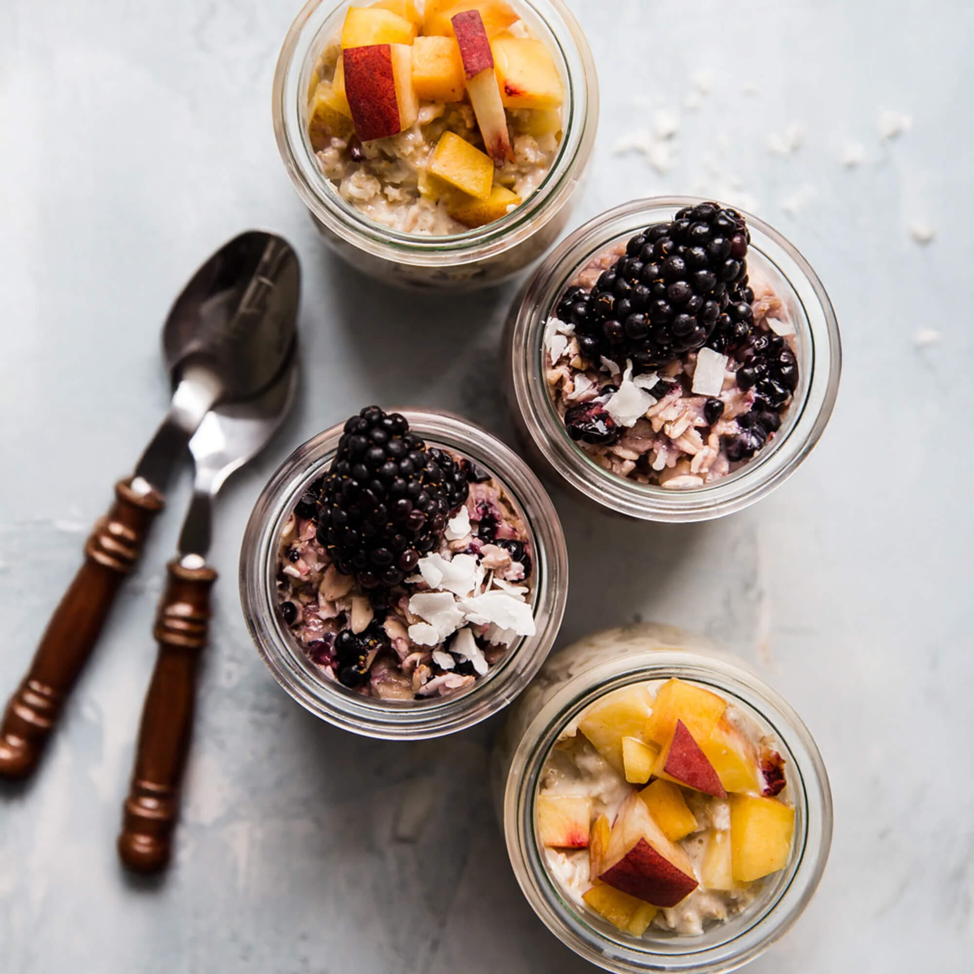 4 glass jars with overnight oats topped with fresh peaches and blackberries on a marble countertop next to 2 spoons