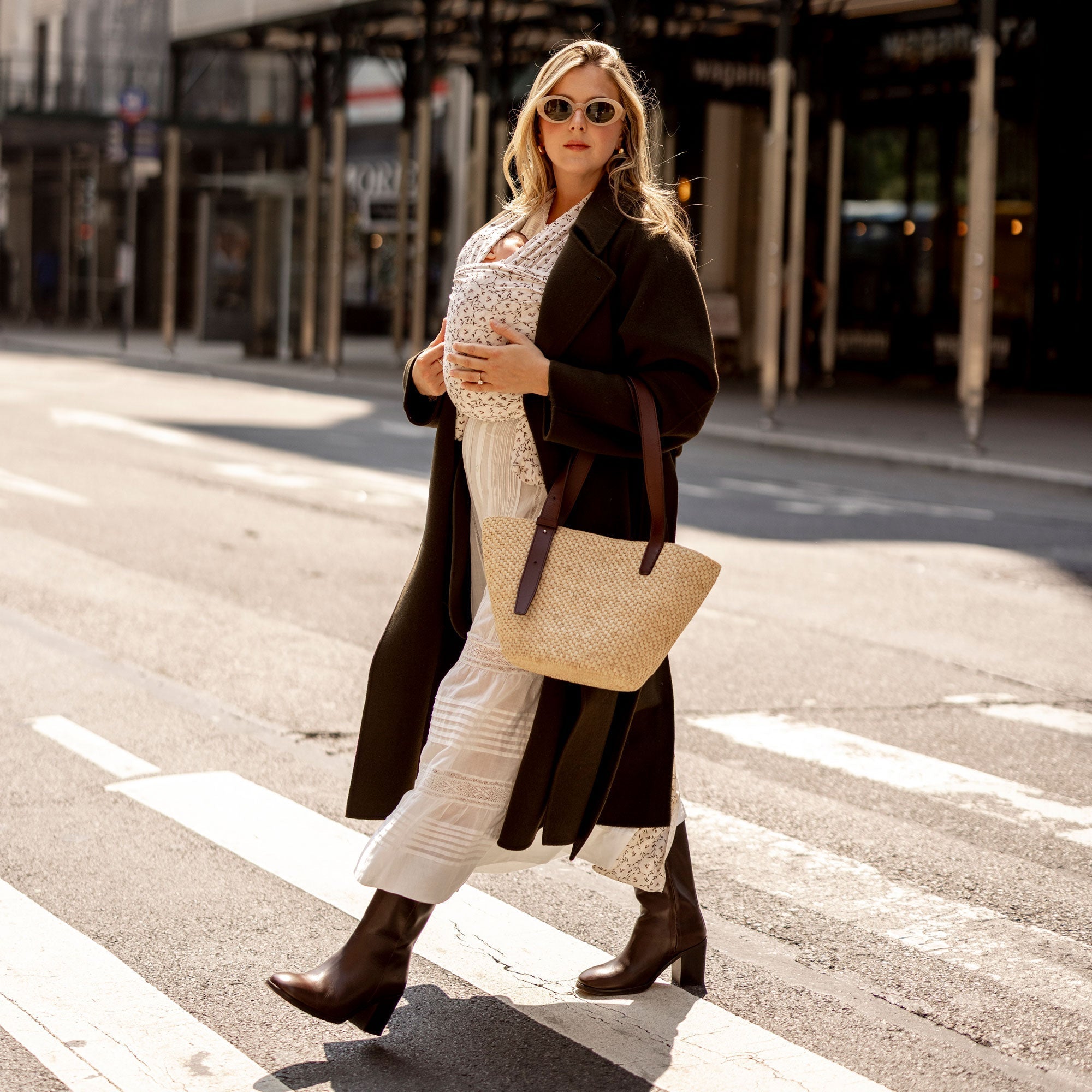 Samantha Varvel walking confidently across a city street wearing the Solly Baby Wrap in Petite Fleurs with her baby nestled inside, paired with a long coat, boots, and a woven tote for a chic, on-the-go motherhood look.