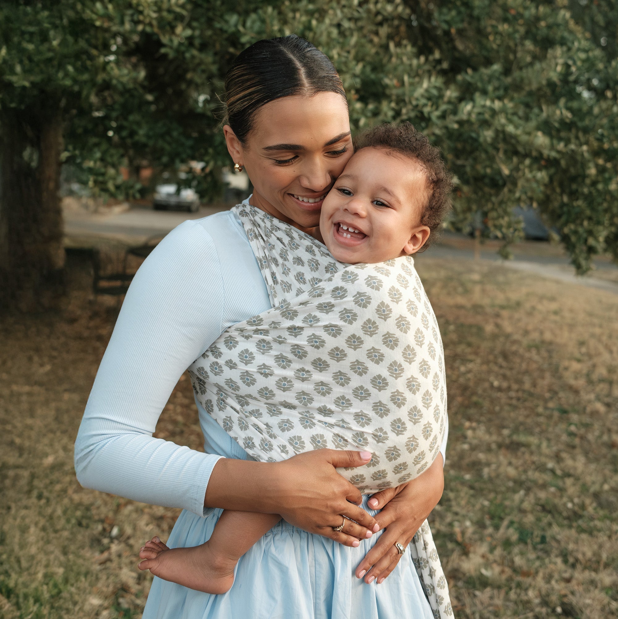 Woman smiles wearing her baby in the Thistle Block print Solly Baby wrap carrier outside in a park
