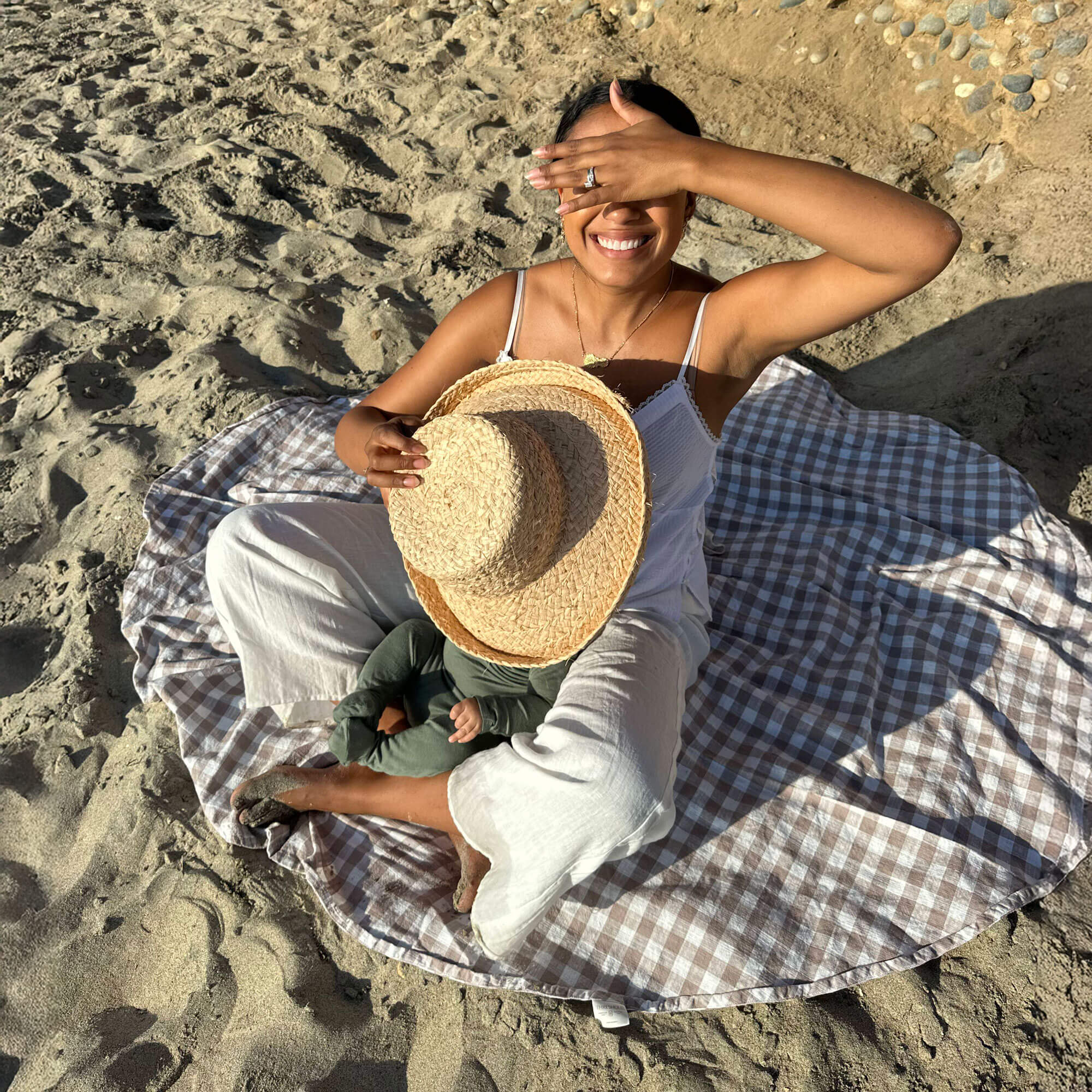 Woman sits on a round gingham blanket on the sand and smiles with one hand shielding her eyes from the sun, and the other holding a straw hat to shield her baby
