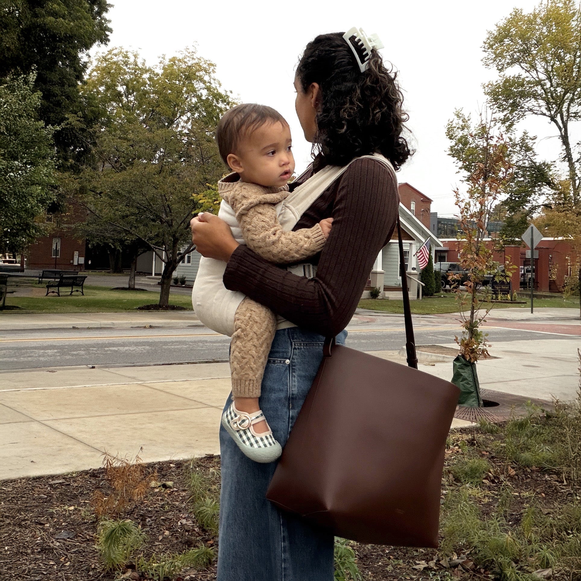 Woman stands in a city square during fall carrying her baby in the Oat Heritage Dot Soft Carrier and with an oversized tote