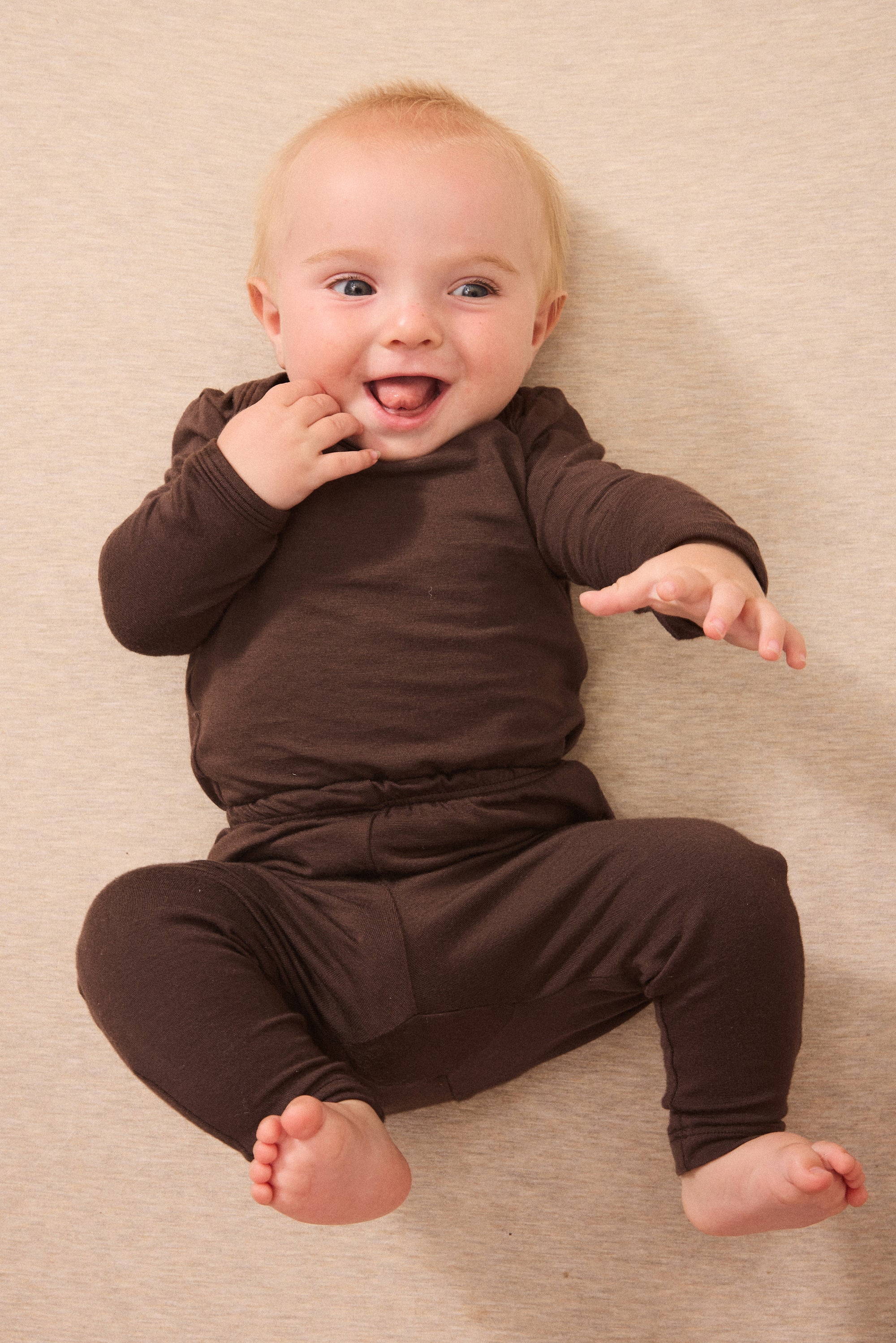 A smiling baby lying on a beige textured blanket, wearing a matching dark brown long-sleeve top and pants set.