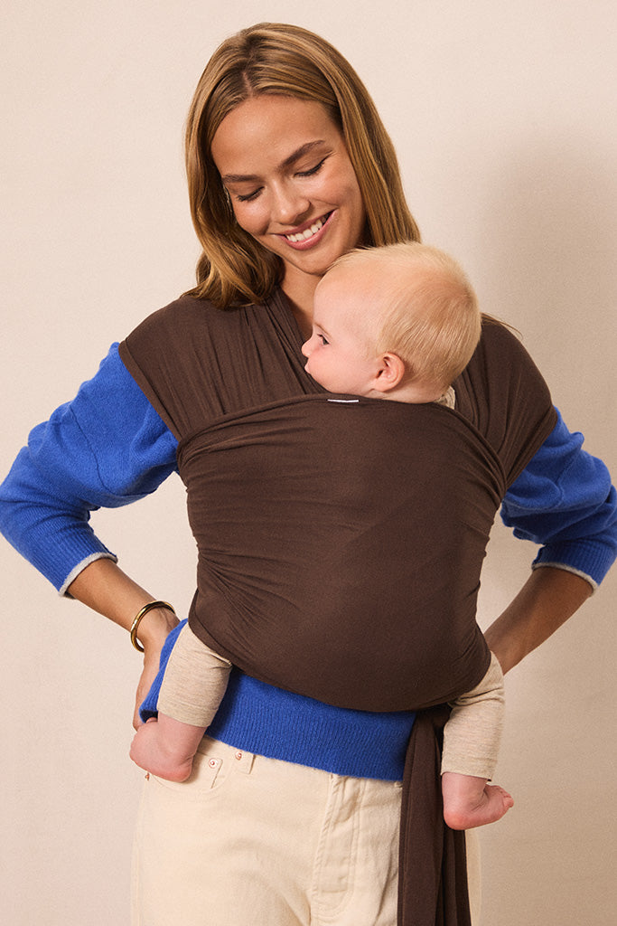 A woman wearing a brown baby wrap carrier with blue long-sleeve shirt underneath, carrying a baby against her chest, set against a plain beige background.