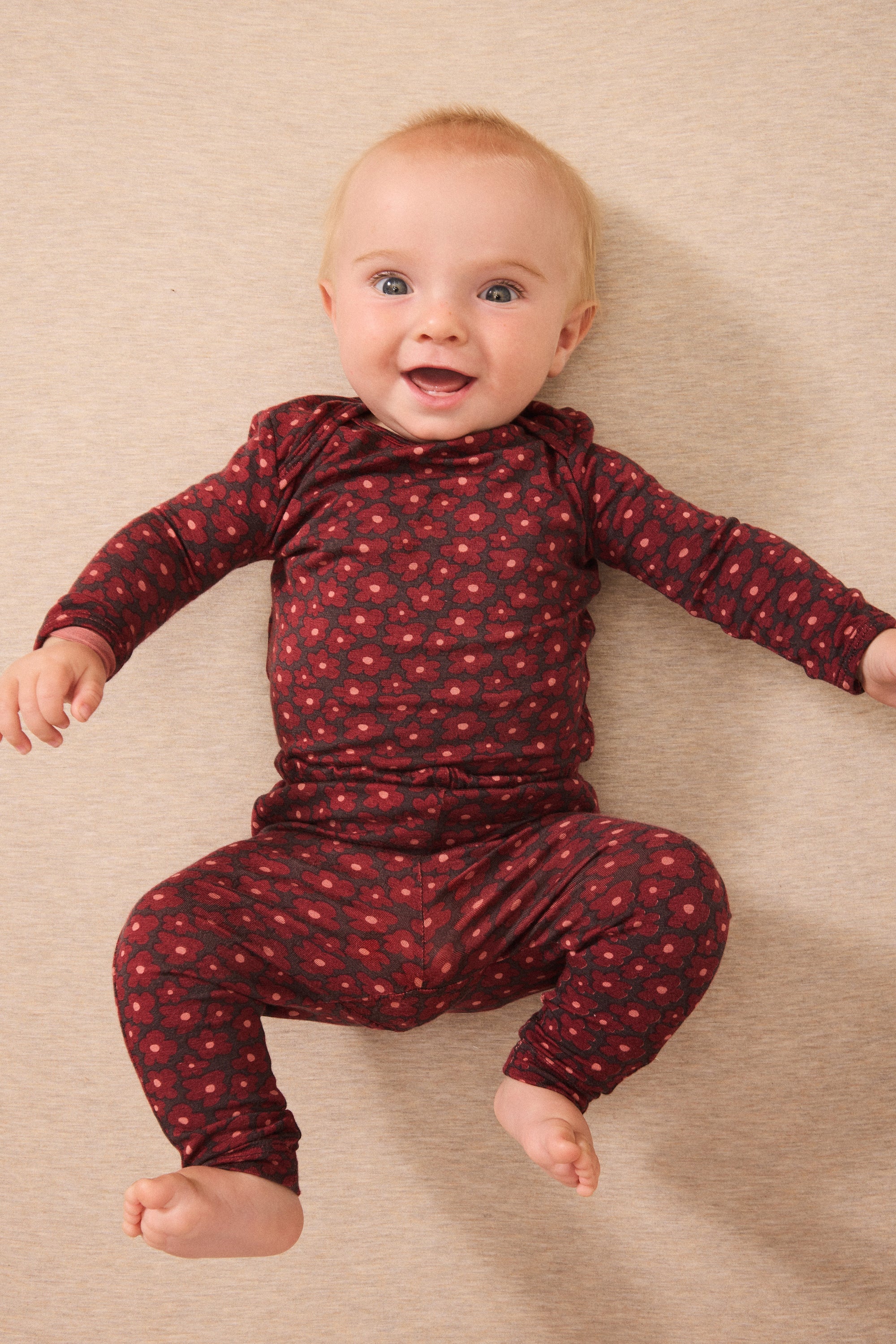A baby wearing a dark red long-sleeve one-piece outfit with a small red floral pattern, lying on a beige textured blanket.