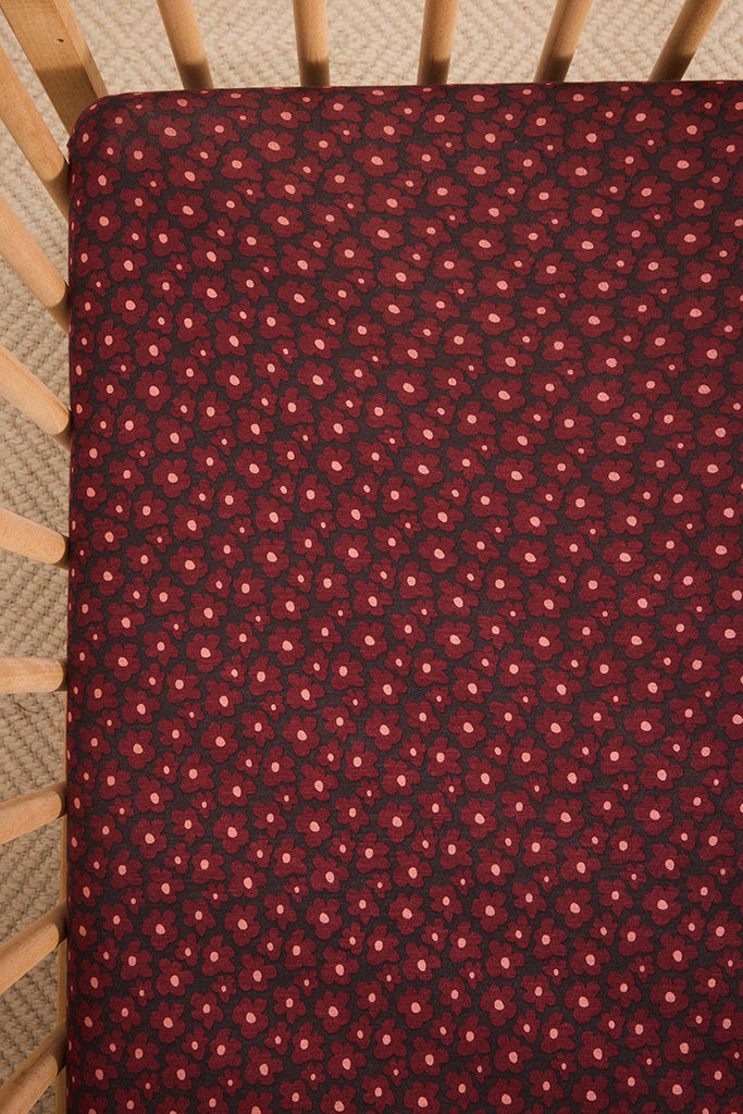 A close-up of a crib mattress fitted sheet with a dark red and black floral pattern, shown inside a wooden crib.