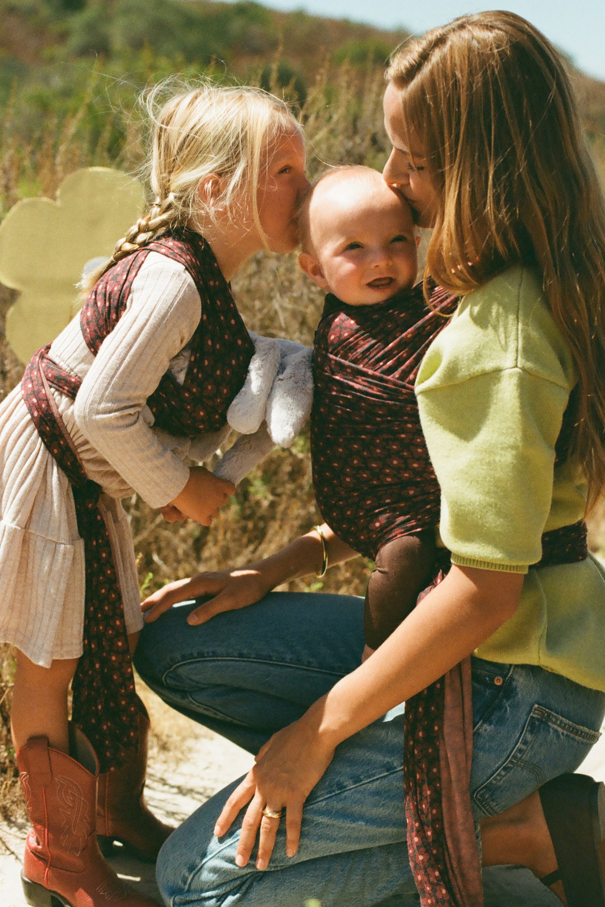 A woman and a young girl outdoors wearing matching dark red patterned baby wraps over their clothing, with the woman also carrying a baby in the wrap. The scene is set in a natural, grassy outdoor environment.