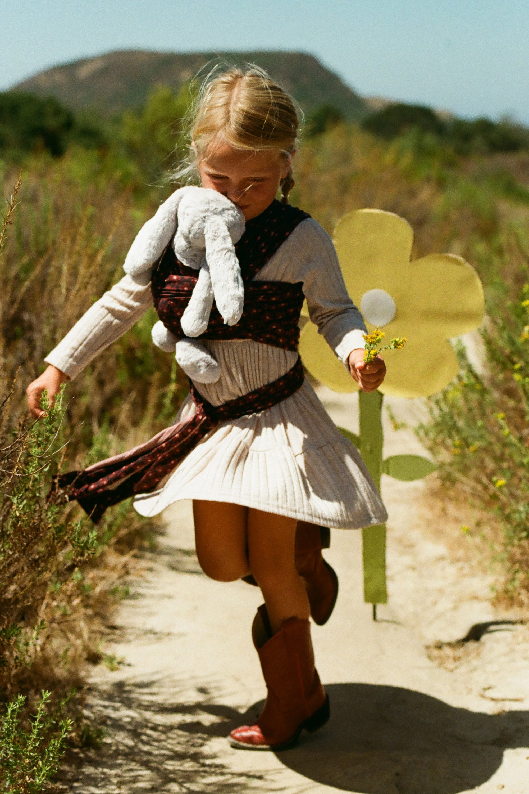 A young girl wearing a cream ribbed dress and brown boots runs along a dirt path outdoors, holding a large yellow flower cutout and a gray stuffed animal.