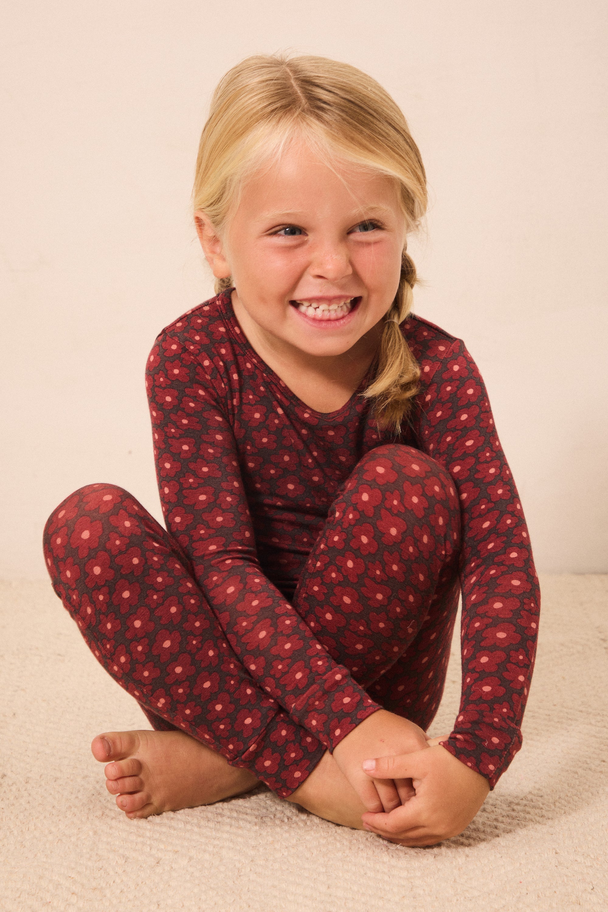 A young blonde girl sitting cross-legged on a light beige carpet, wearing a long-sleeve red pajama top and matching leggings with a small repeating floral pattern.