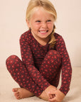 A young blonde girl sitting cross-legged on a light beige carpet, wearing a long-sleeve red pajama top and matching leggings with a small repeating floral pattern.