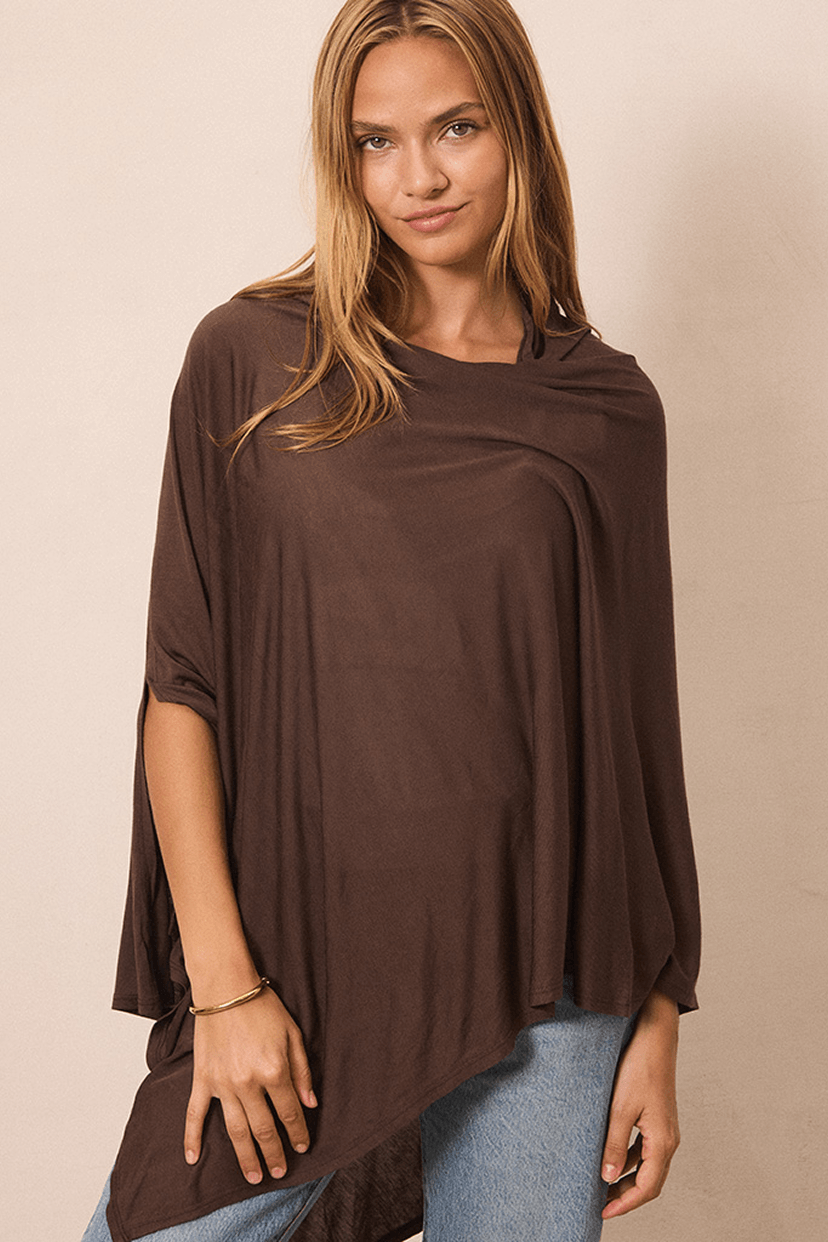 A woman wearing a draped, oversized brown top with three-quarter sleeves and an asymmetrical hem, paired with light blue jeans, against a plain beige background.