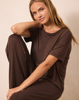 A woman wearing a matching dark brown short-sleeve t-shirt and wide-leg pants, sitting on a stool against a plain beige studio background.