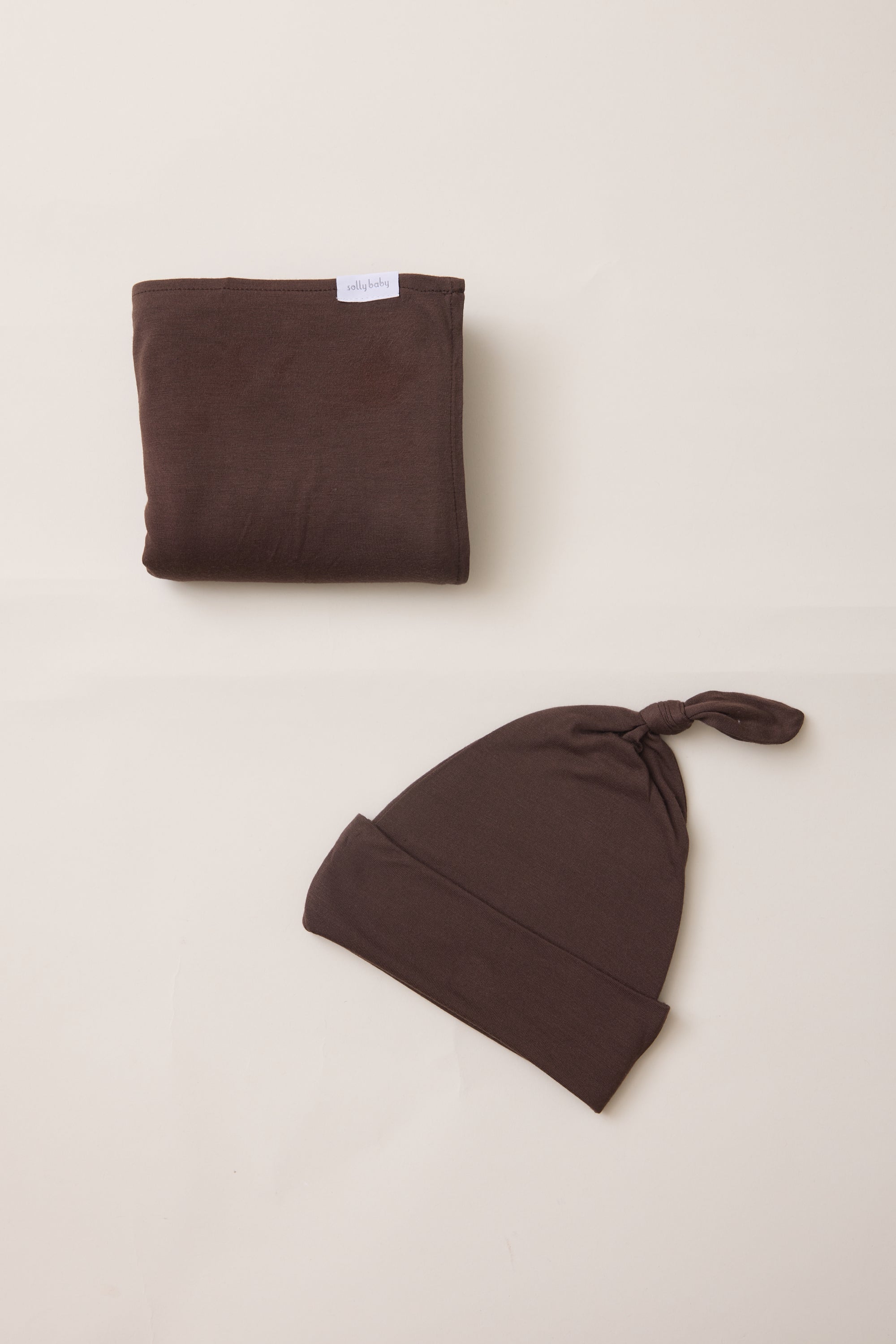 A folded dark brown fabric item with a small white tag is placed above a matching dark brown knotted beanie on a plain warm off-white background.