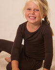 A young blonde girl smiling while wearing a dark brown long-sleeve top and matching dark brown pants, sitting on a light beige carpet against a plain beige wall.