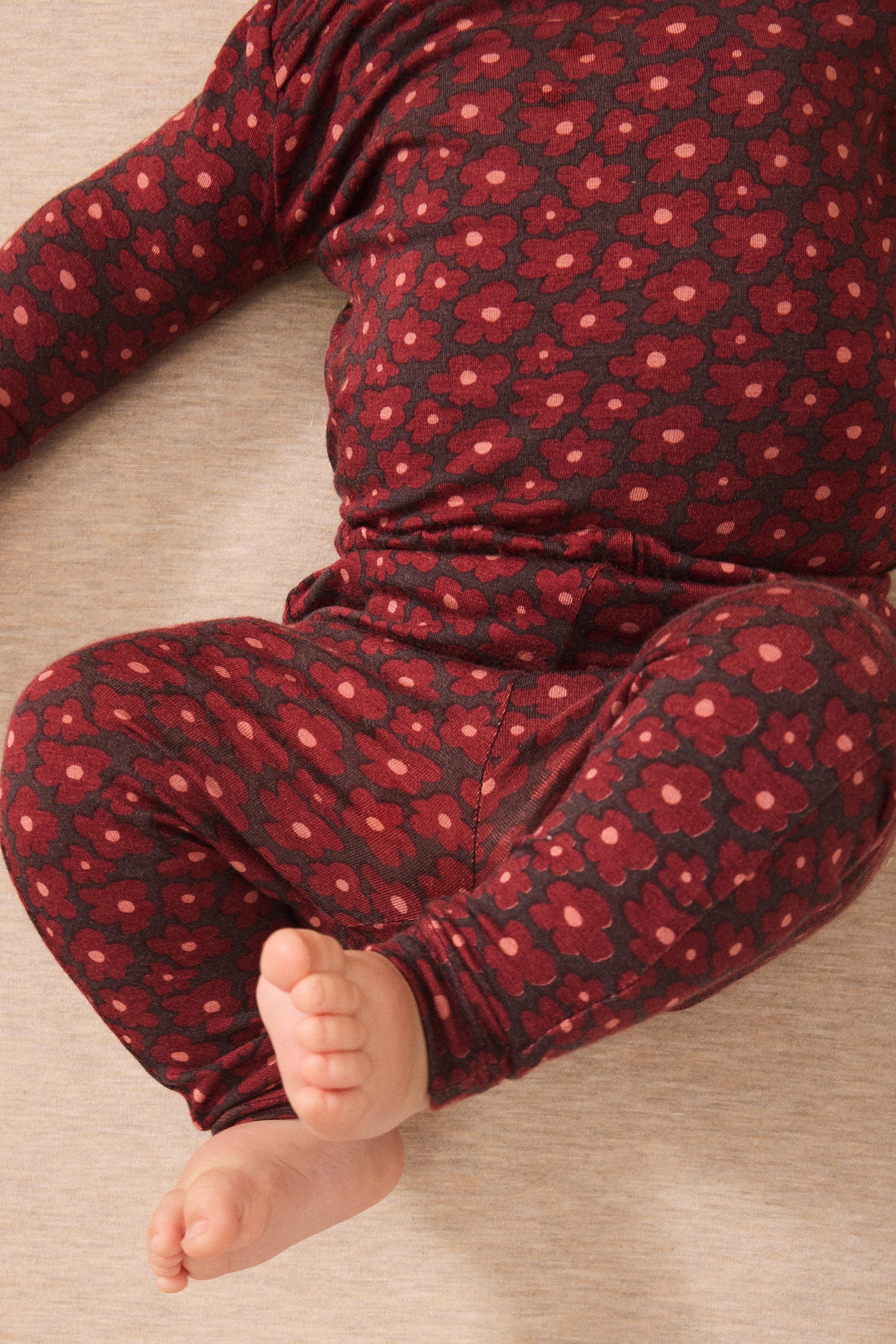 A close-up of a baby wearing a dark red and black floral patterned long-sleeve pajama set, lying on a light beige textured floor.