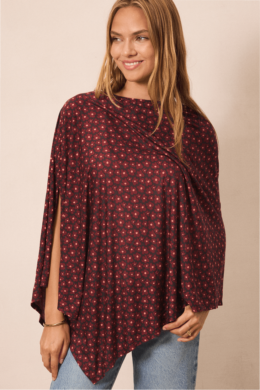 A woman wearing a dark pink flower patterned poncho top with a small white and red floral pattern, paired with light blue jeans, against a solid beige background.