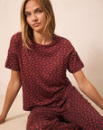 A woman wearing a matching short-sleeve t-shirt and wide-leg pants pajama set in a deep red color with a small repeating yellow floral pattern, sitting against a plain beige background.