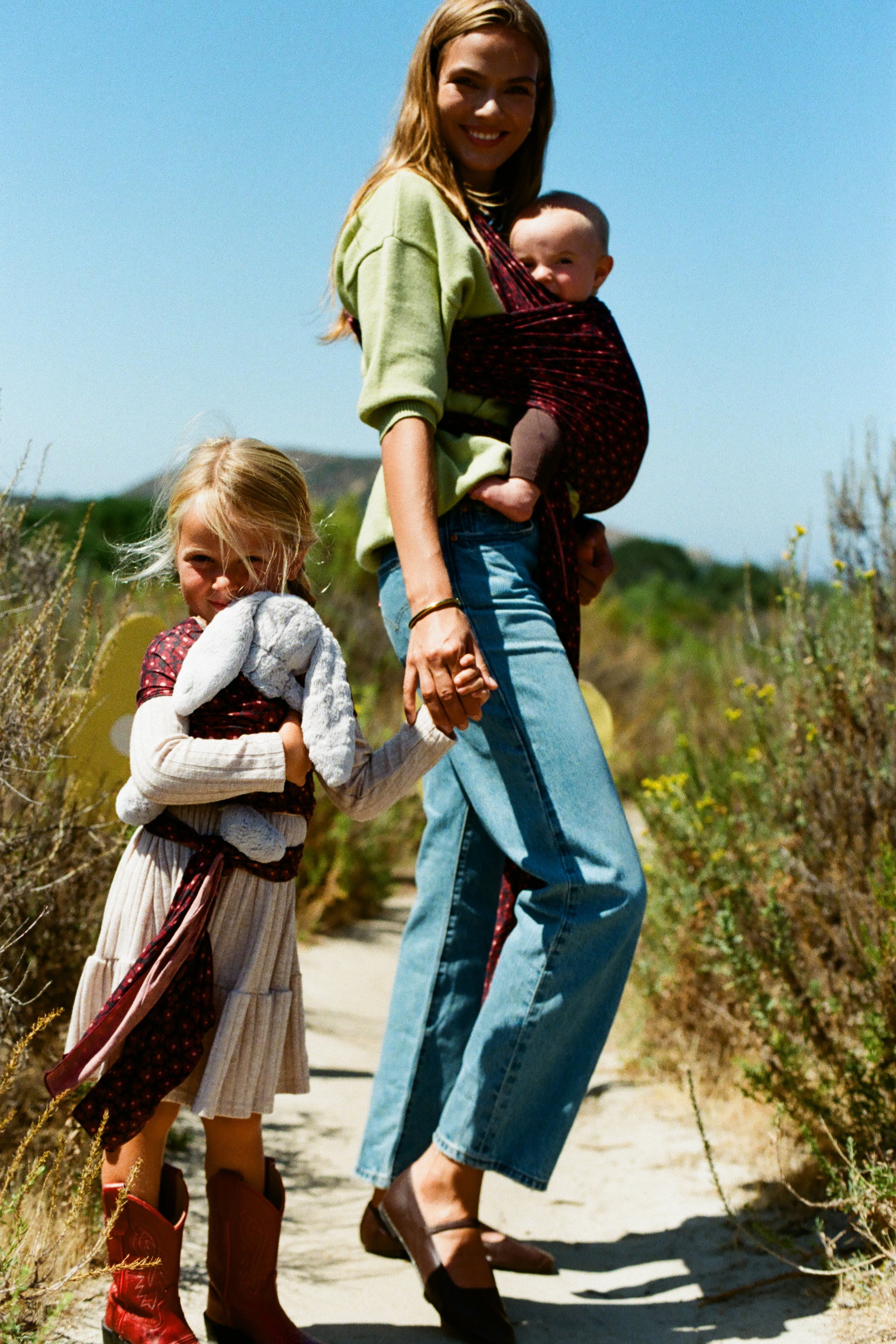 A woman carrying a baby in a dark red patterned baby carrier wrap walks outdoors on a dirt path with a young girl beside her holding a gray stuffed bunny.